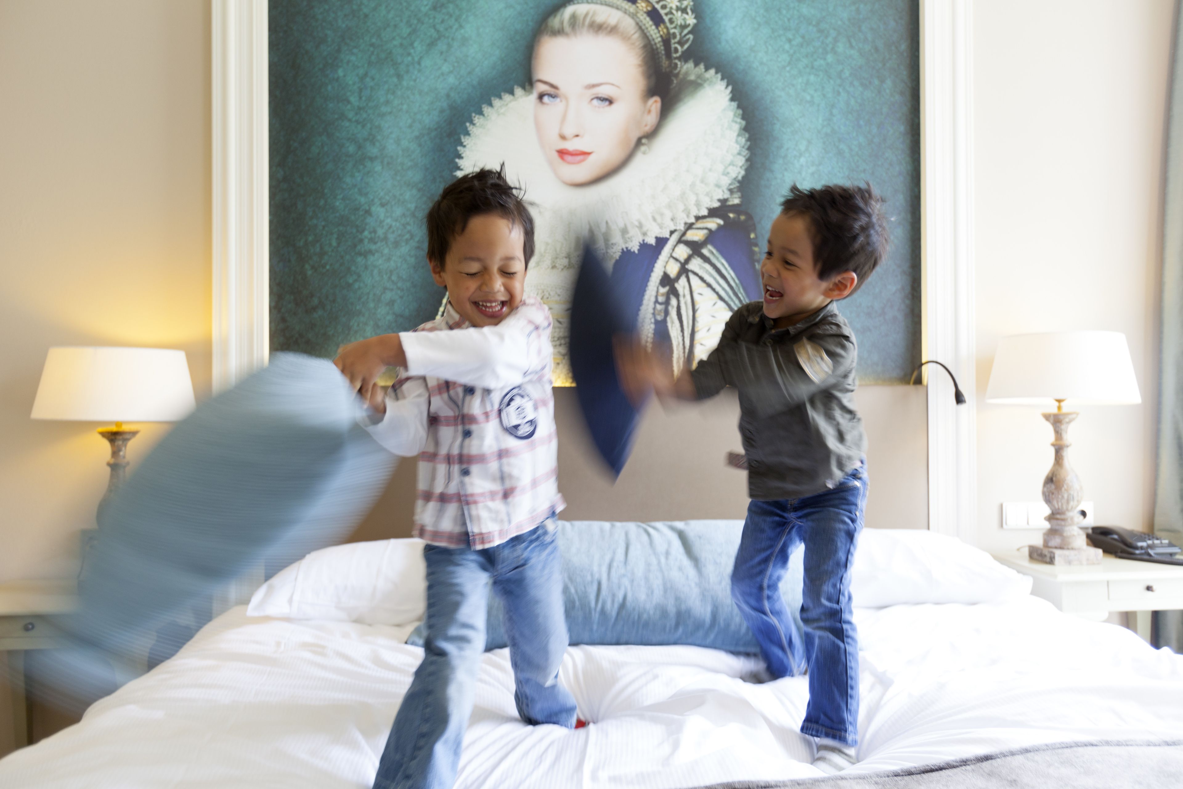 Twee kinderen spelen op een bed in een hotelkamer, met een groot portret aan de muur en lampen aan weerszijden.