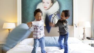 Twee kinderen spelen op een bed in een hotelkamer, met een groot portret aan de muur en lampen aan weerszijden.