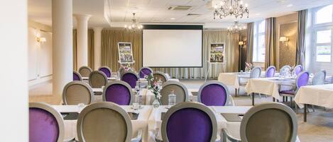 A conference room with tables and chairs, a projector screen, and a chandelier.