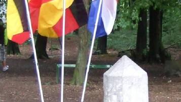 Eine Fahnenmast mit mehreren Flaggen, ein Steinmonument und ein bewaldeter Hintergrund.