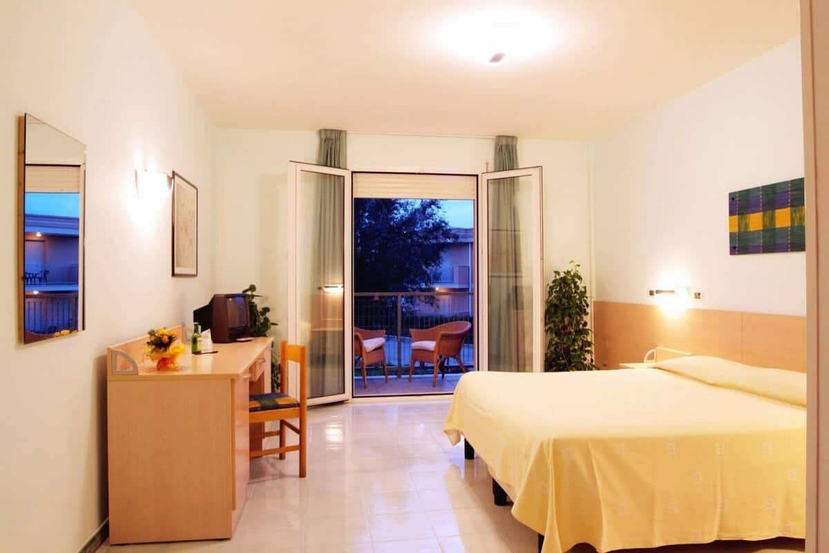 A hotel room with a bed, a desk with a chair, a television, and a balcony with chairs.