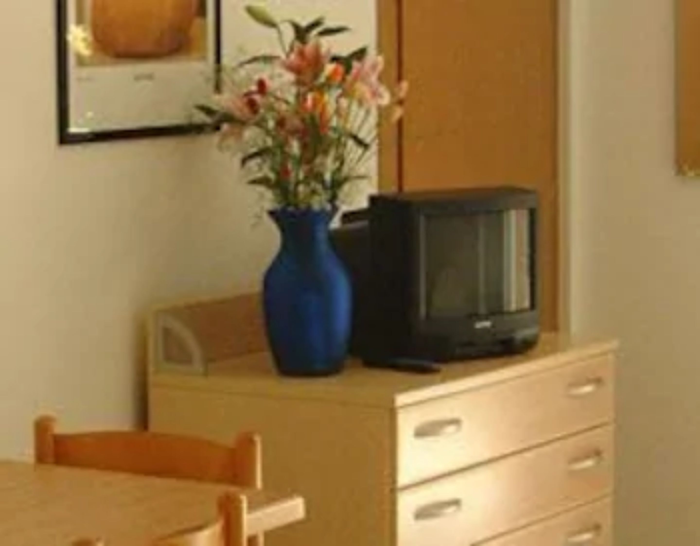A room with a wooden dresser, a blue vase with flowers, and a television on top.