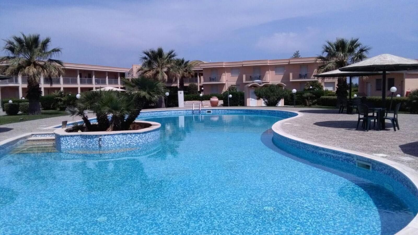 A swimming pool with a curved design, surrounded by palm trees and a building with balconies in the background.