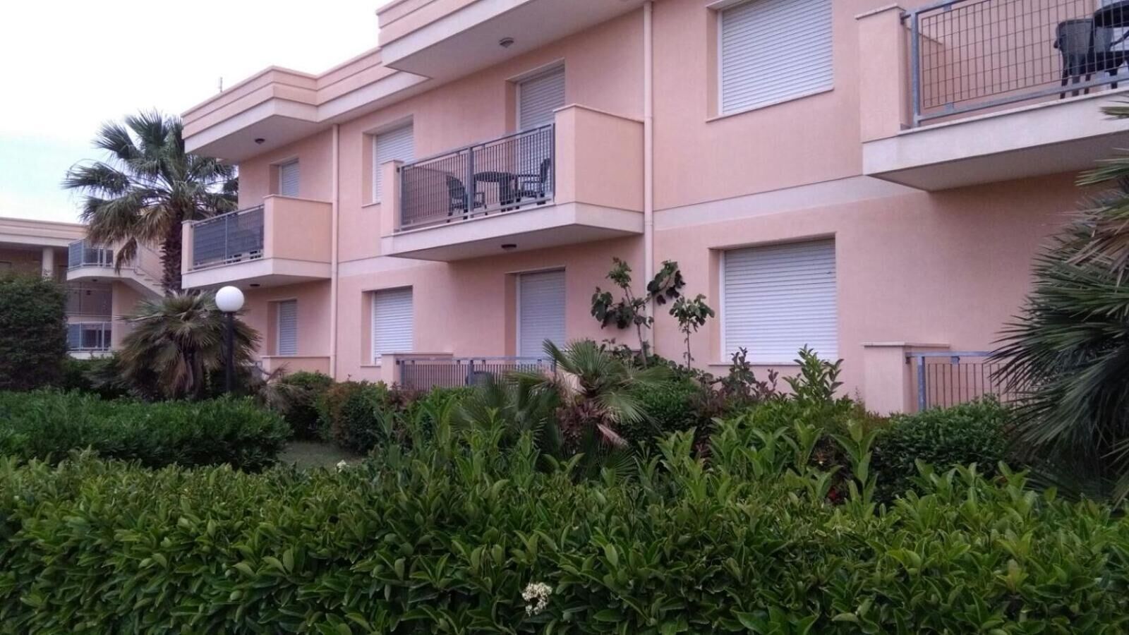 A multi-story residential building with balconies, a well-maintained garden, and a palm tree.