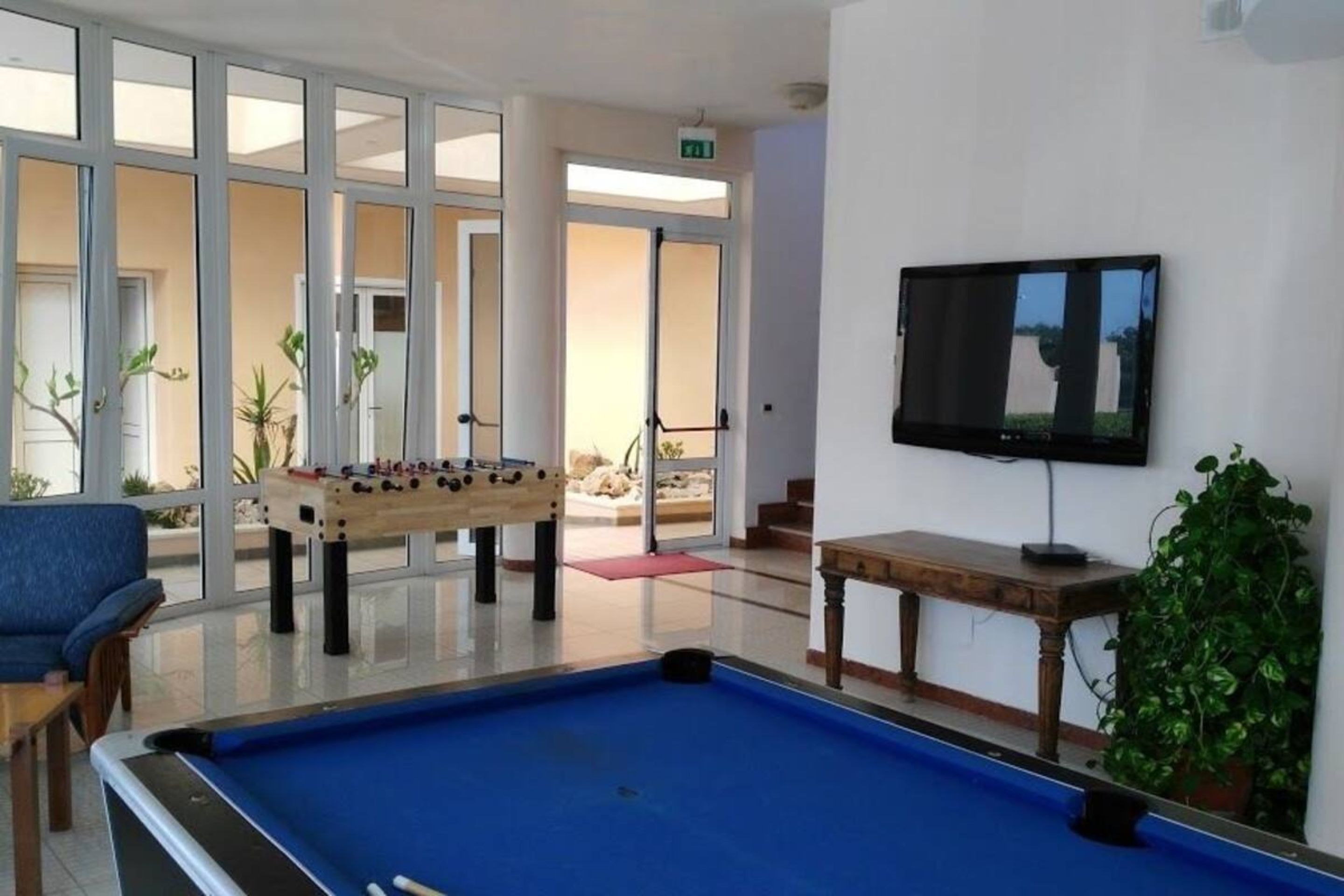 Una mesa de billar, una mesa de fútbol de mesa y una televisión en una habitación con ventanas amplias y una planta.