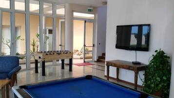 Una mesa de billar, una mesa de fútbol de mesa y una televisión en una habitación con ventanas amplias y una planta.