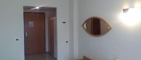 Quarto de hotel com porta de madeira, espelho na parede e uma pequena mesa com cadeira.