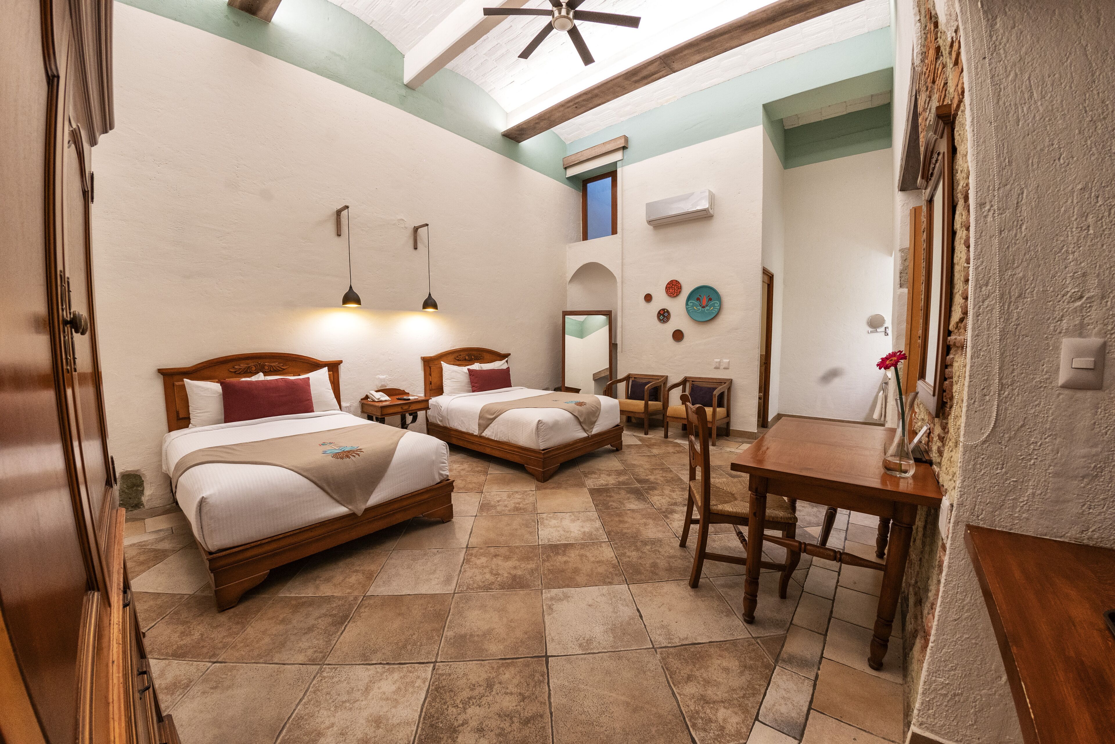 A hotel room with two beds, a desk, and a chair. There is a ceiling fan, a mirror, and a small table with a vase.