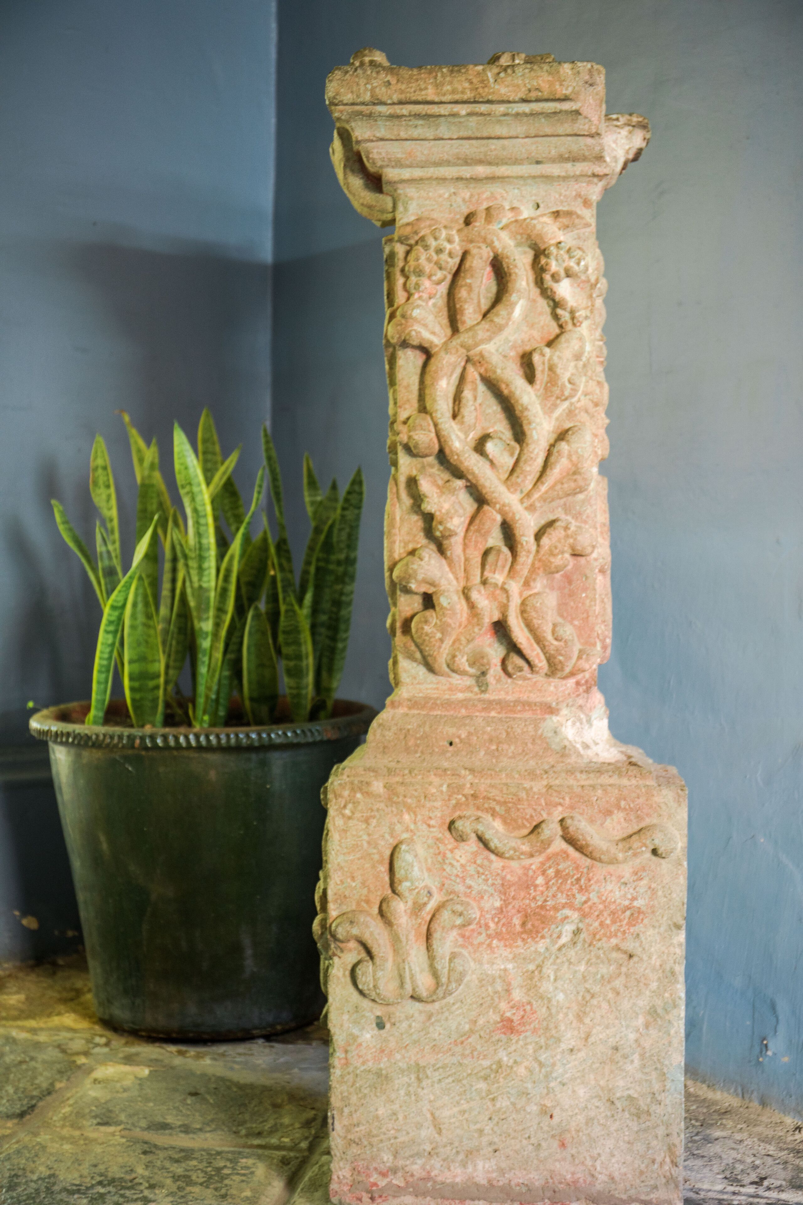 A stone column with intricate carvings, a potted plant, and a plain wall.