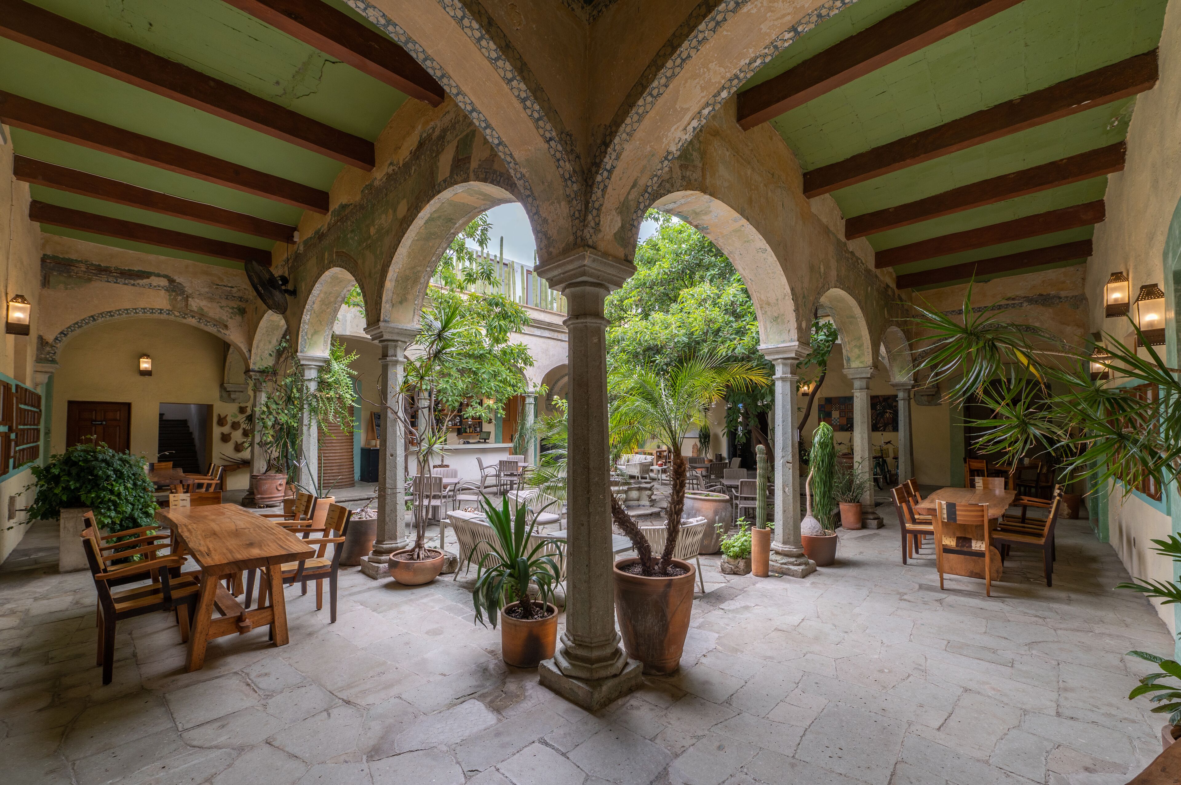 Une cour intérieure aménagée avec des tables et des chaises en bois, des plantes en pot et des arcades.