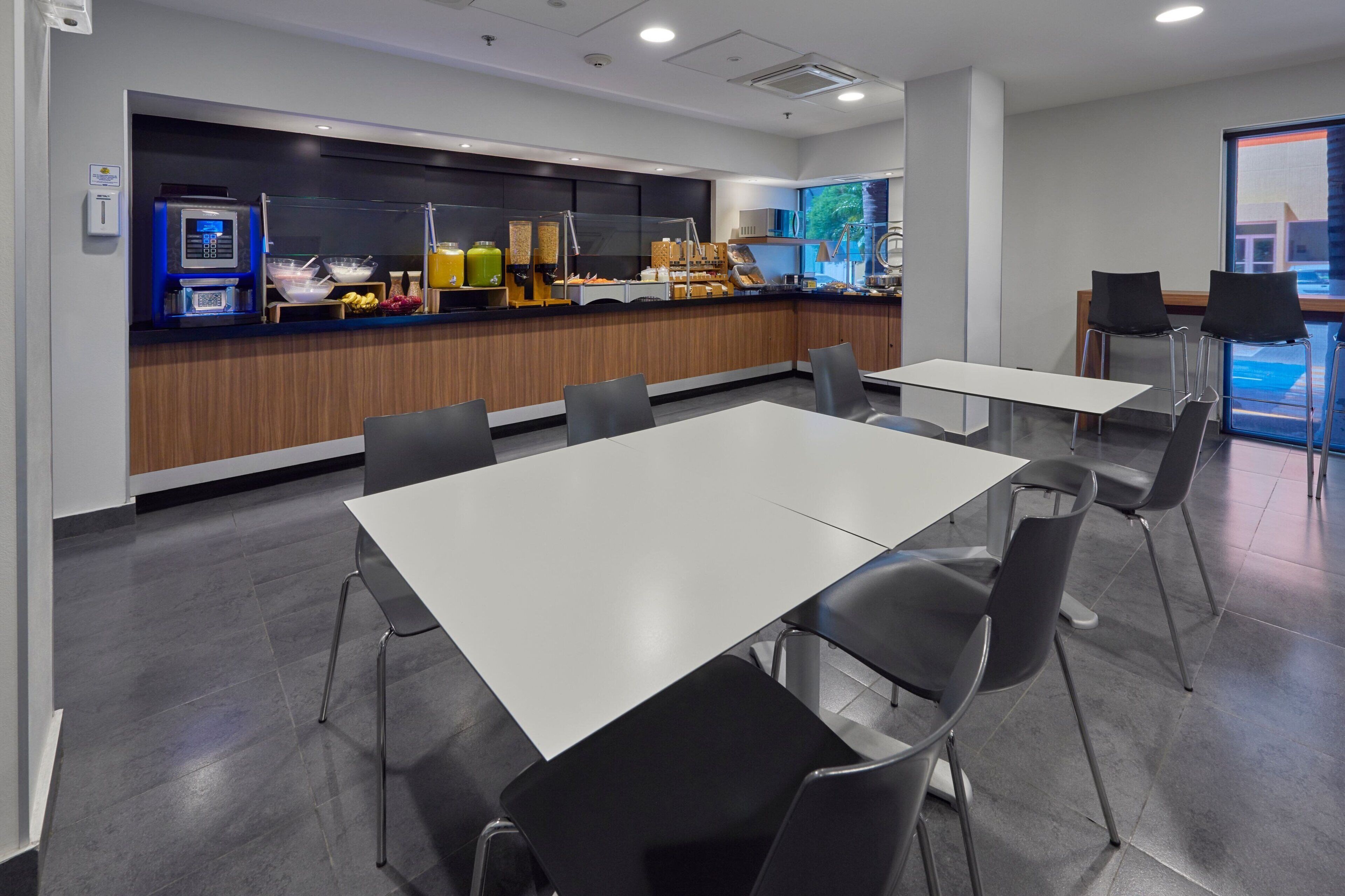 A modern dining area with a self-service counter, white tables, and black chairs.