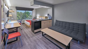 Ein modernes Hotelzimmer mit einem Bett, einem Schreibtisch, einem Sessel, einer kleinen Küchenzeile und einem großen Fenster mit Blick ins Freie.