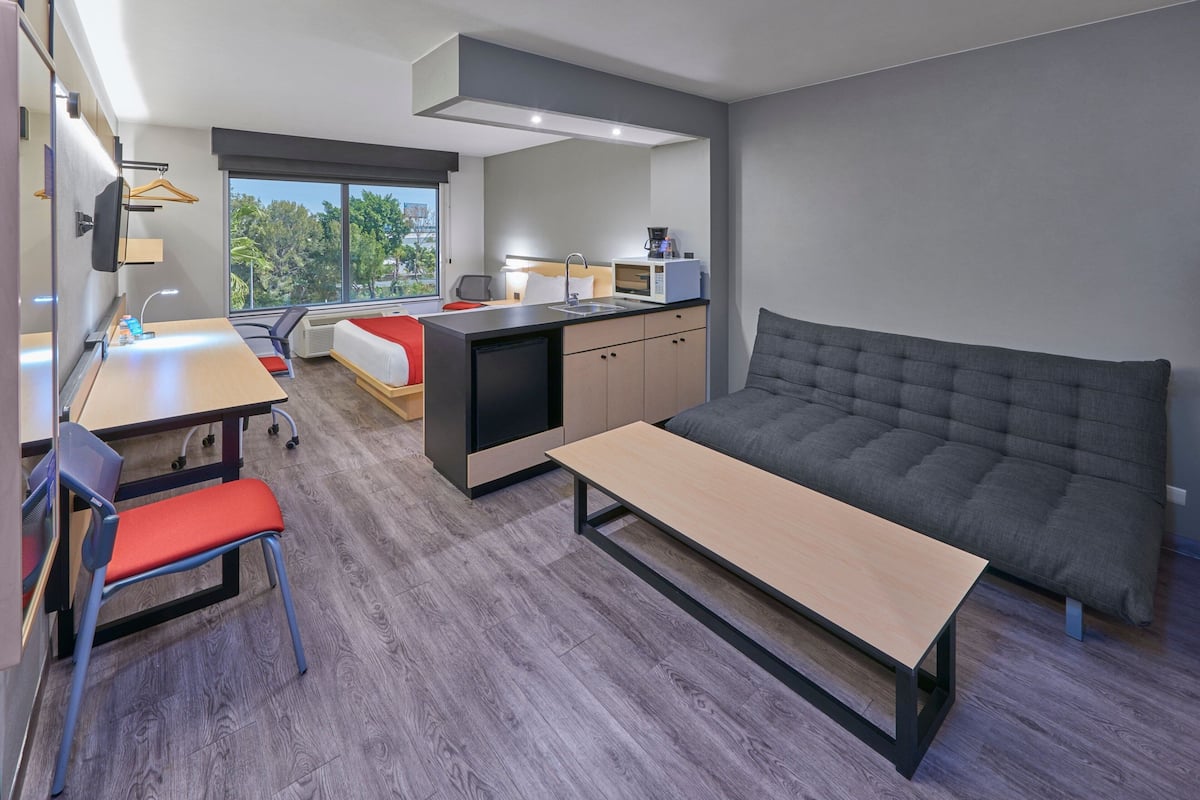 A modern hotel room with a bed, a desk, a chair, a small kitchenette, and a large window offering a view of the outdoors.