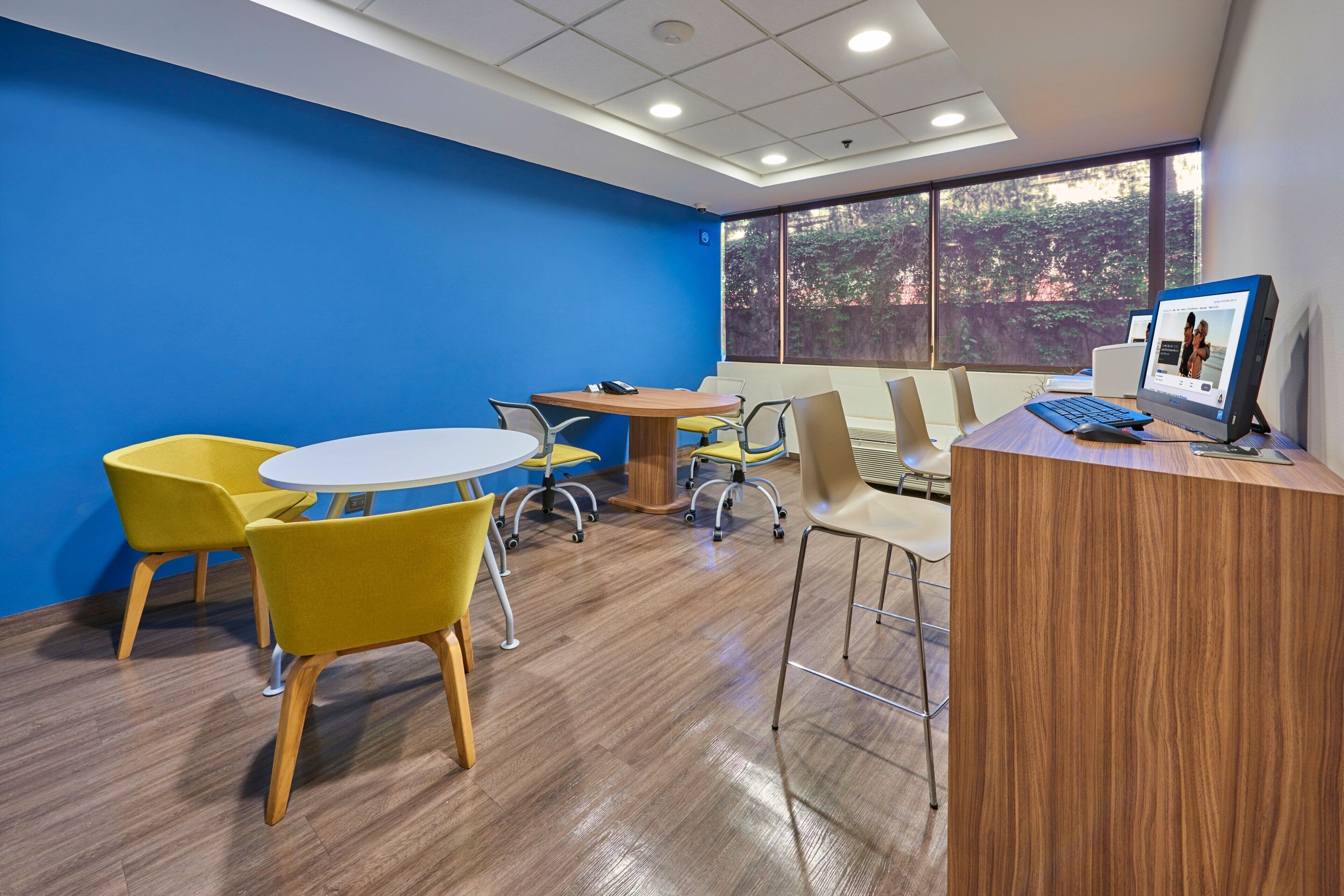 A conference room with a round table, yellow chairs, a wooden desk with a computer, and large windows with a view of trees.