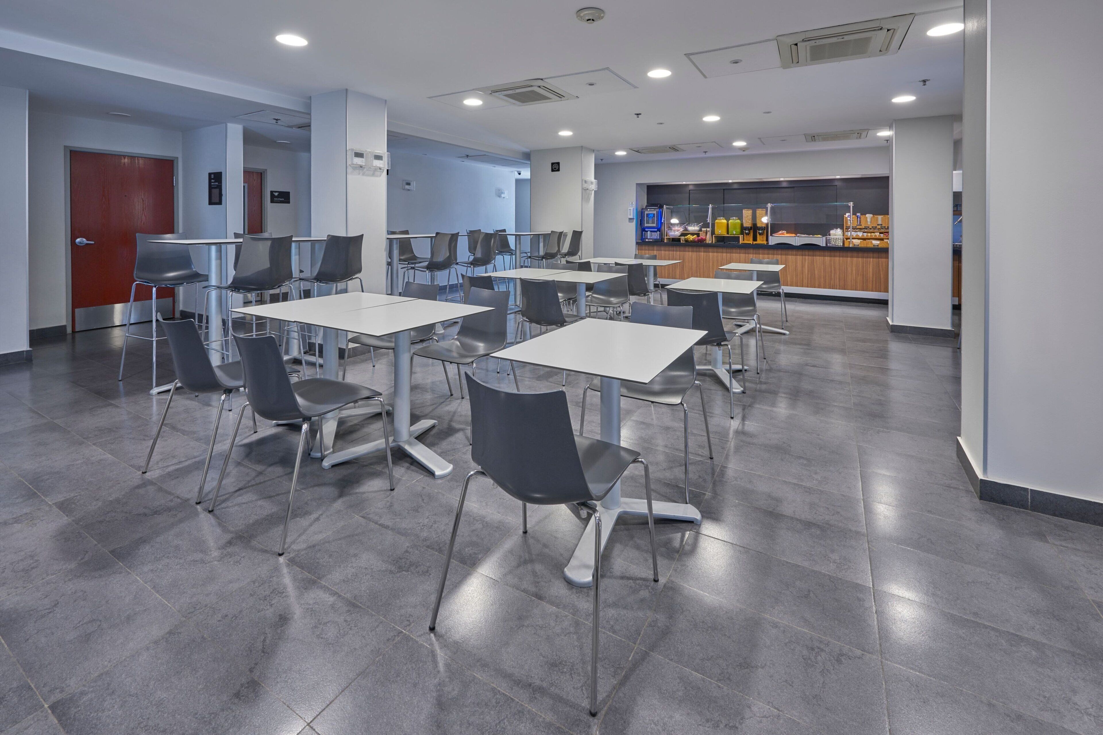 A modern cafeteria with tables and chairs, a counter with food items, and a door in the background.