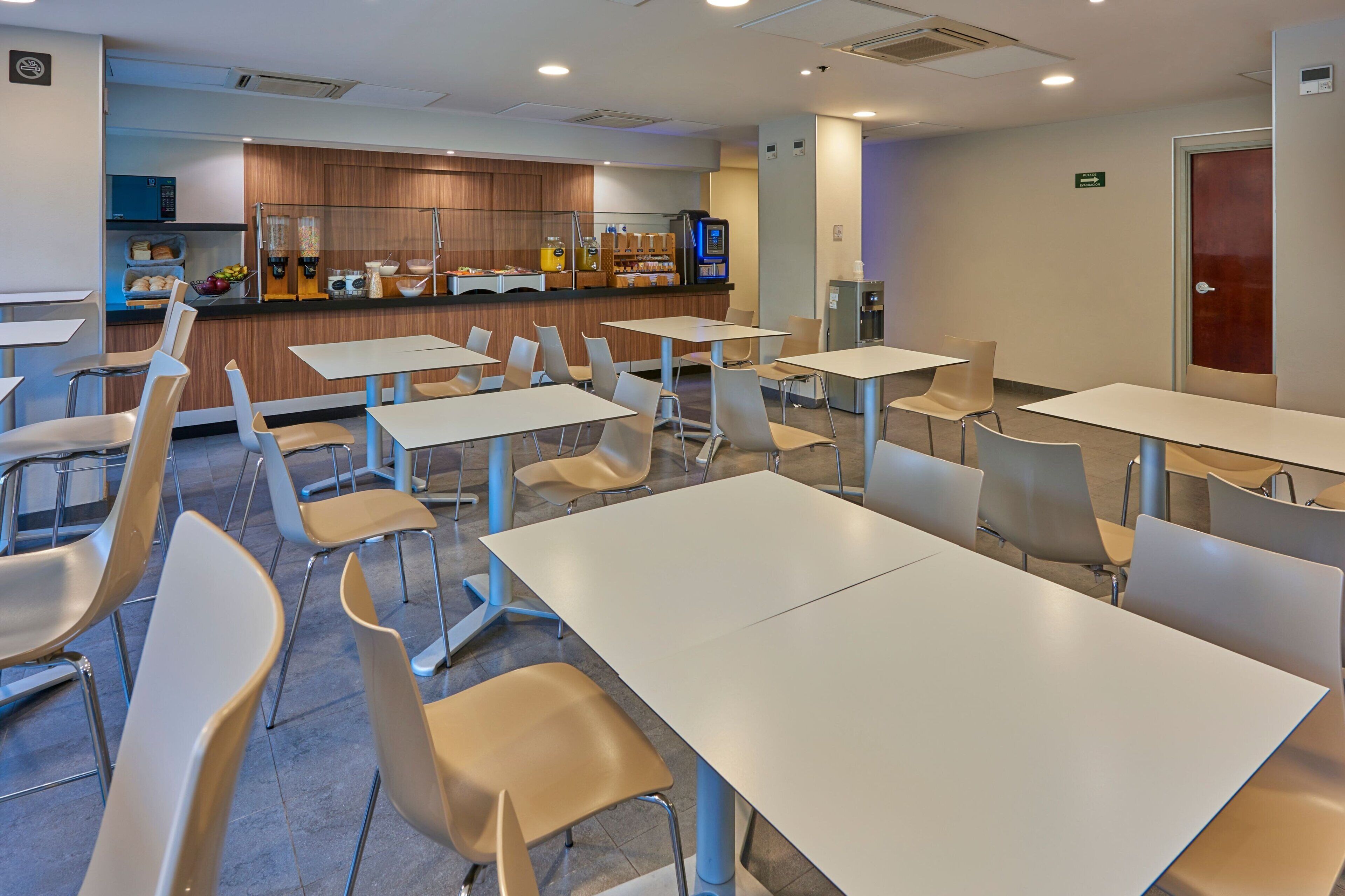 Una cafetería moderna con mesas y sillas blancas, una zona de cocina con una máquina de café y un dispensador de agua.