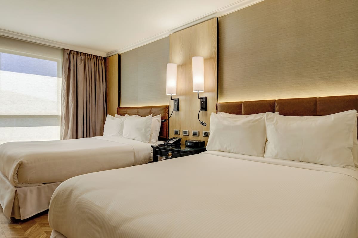 A hotel room with two beds, a nightstand with a phone, and a wall-mounted lamp.