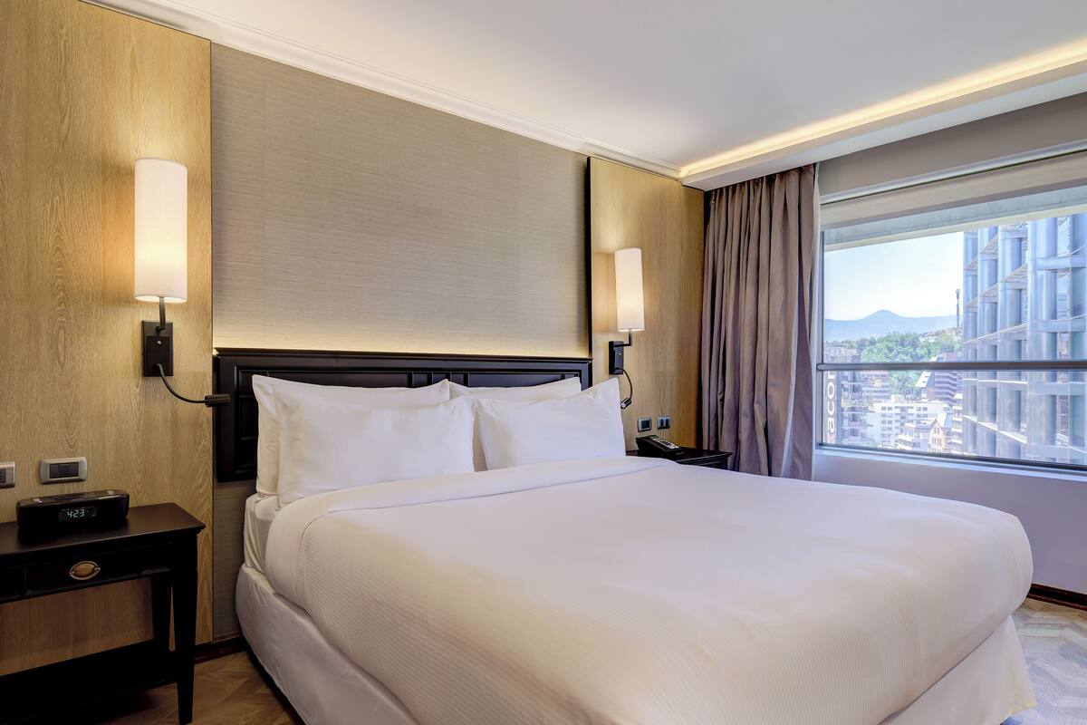 A hotel room with a large bed, two bedside tables, a nightstand, a wall-mounted lamp, and a view of the city through a large window.