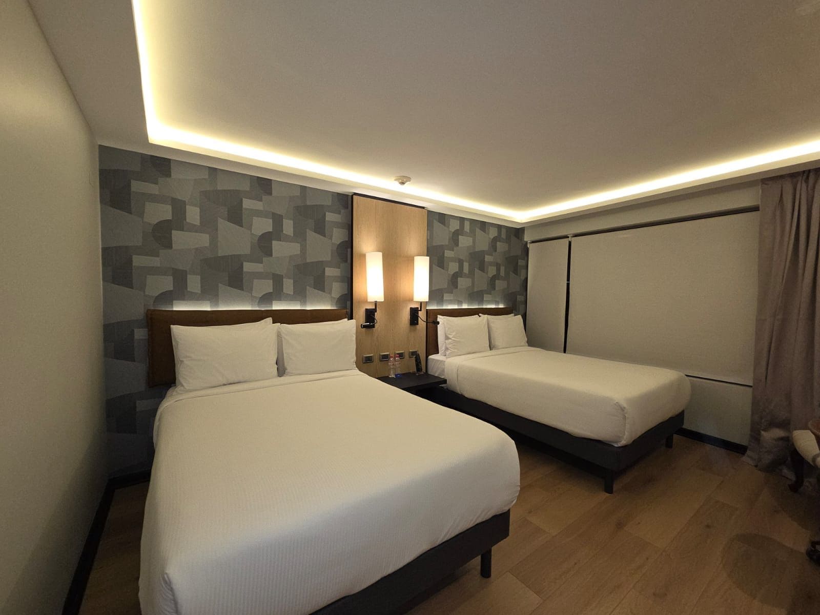 Een hotelkamer met twee bedden, een nachtkastje, een wandlamp en een raam met gordijnen.