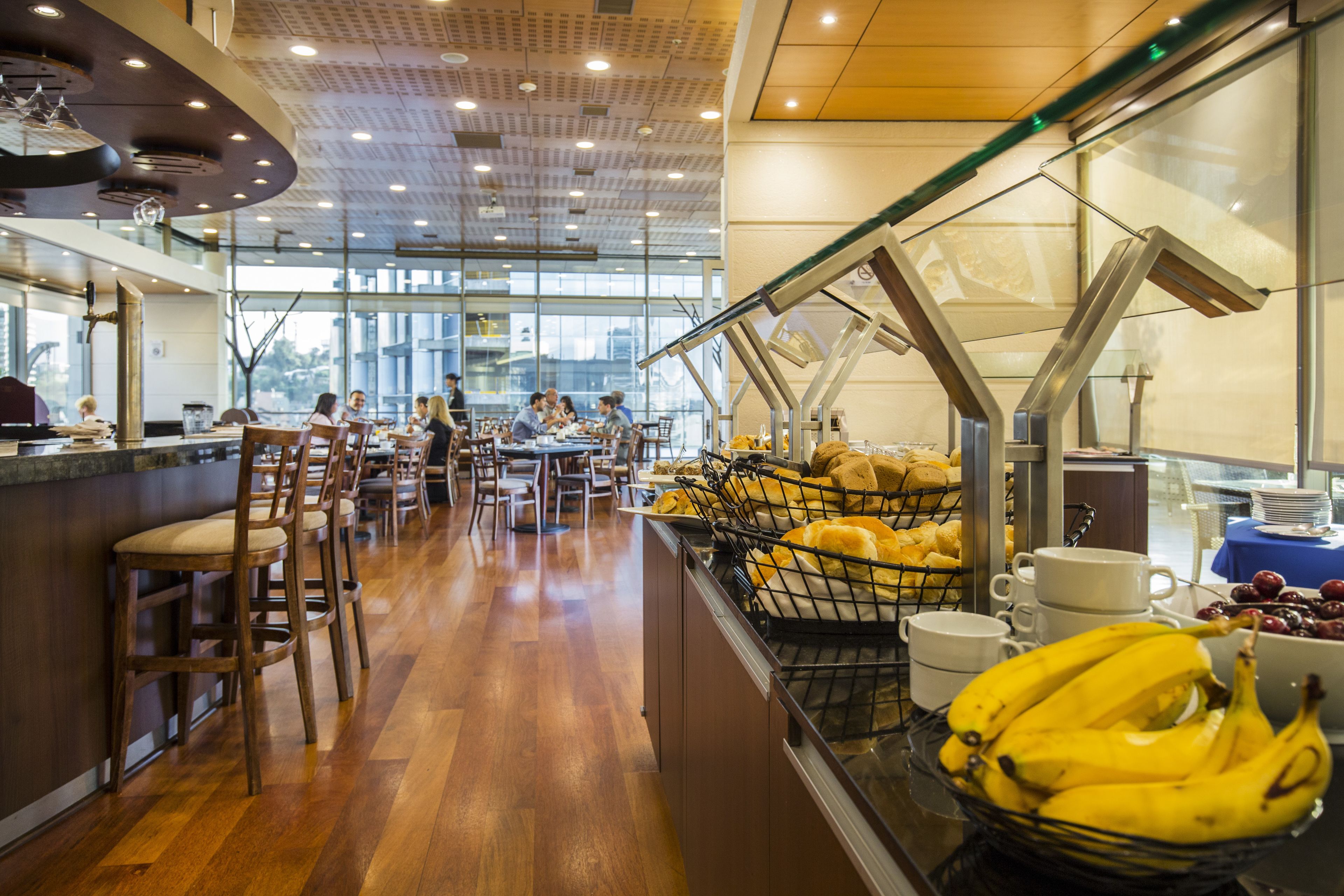 A modern restaurant with wooden flooring, a glass partition, and a buffet area with fruits and bread.