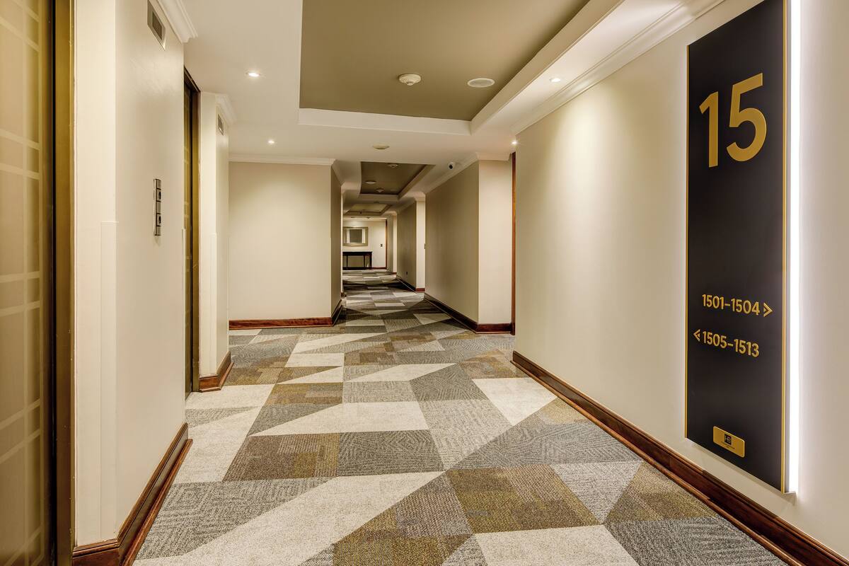 A hotel hallway with a geometric patterned carpet, a number 15 sign on the wall, and a view into a room at the end.