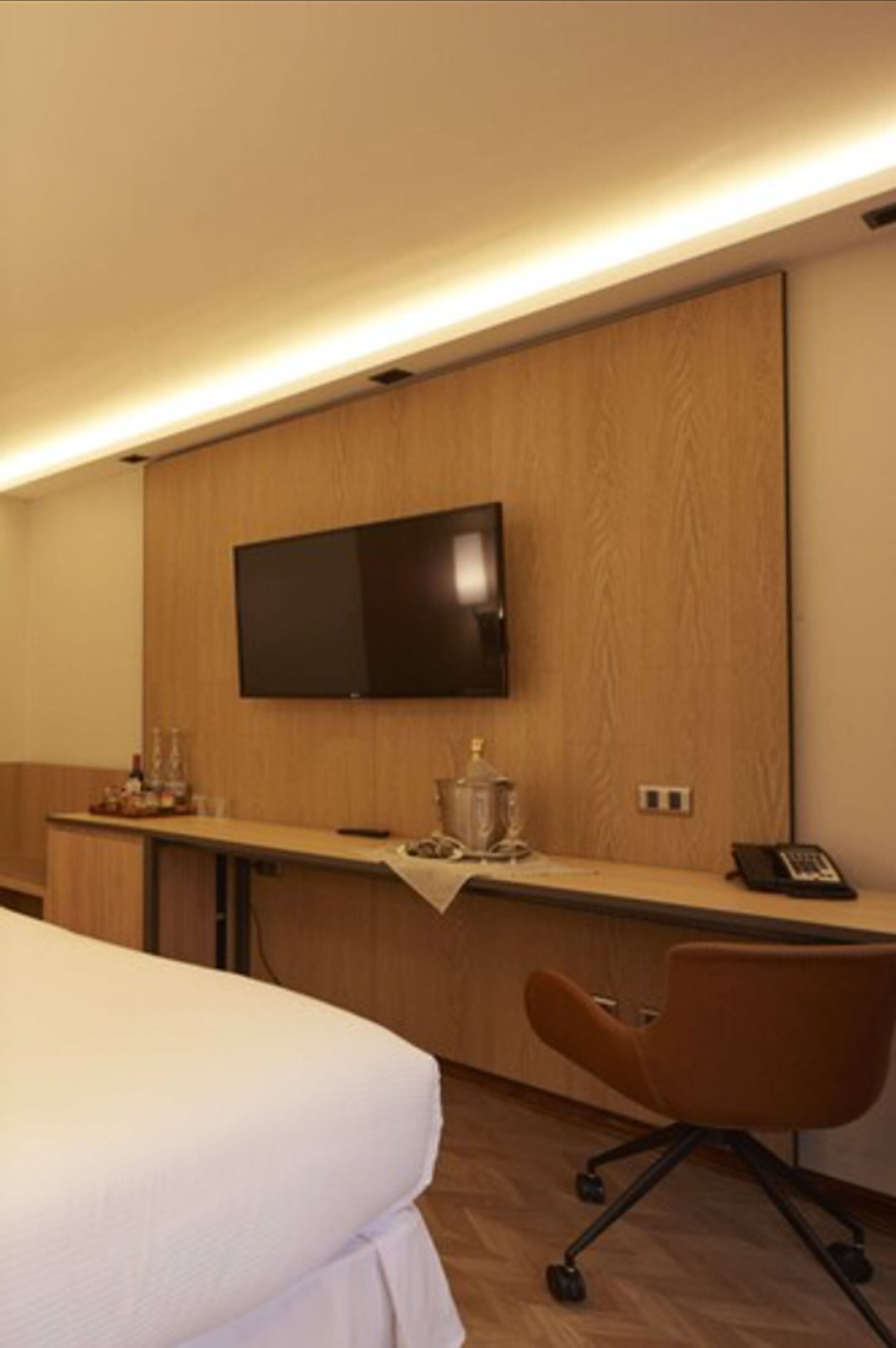 A hotel room with a bed, a desk, a chair, a television mounted on the wall, and a small table with a bottle and glasses.