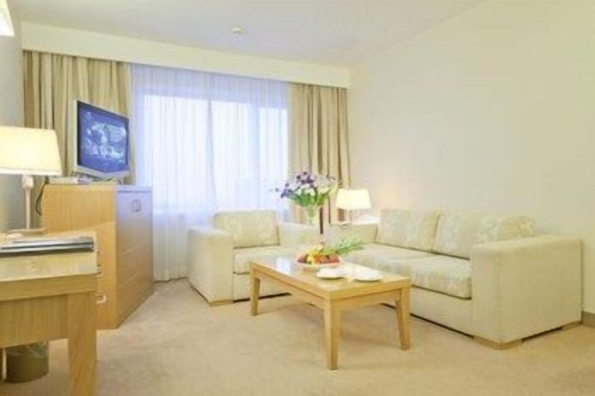 Une chambre d’hôtel avec un canapé, une table basse ornée d’un vase et de fruits, une télévision et un bureau avec une lampe.