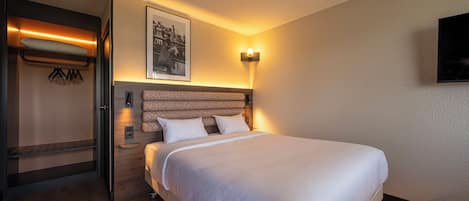 A hotel room with a bed, a headboard, a wall-mounted light, a framed picture, and a coat rack with hangers.