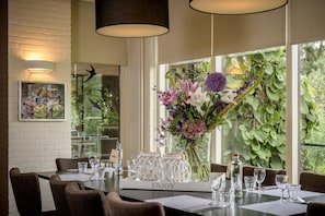 Spiseplads med et bord dækket til seks personer, en dekorativ vase med blomster og et billede hængt på væggen.