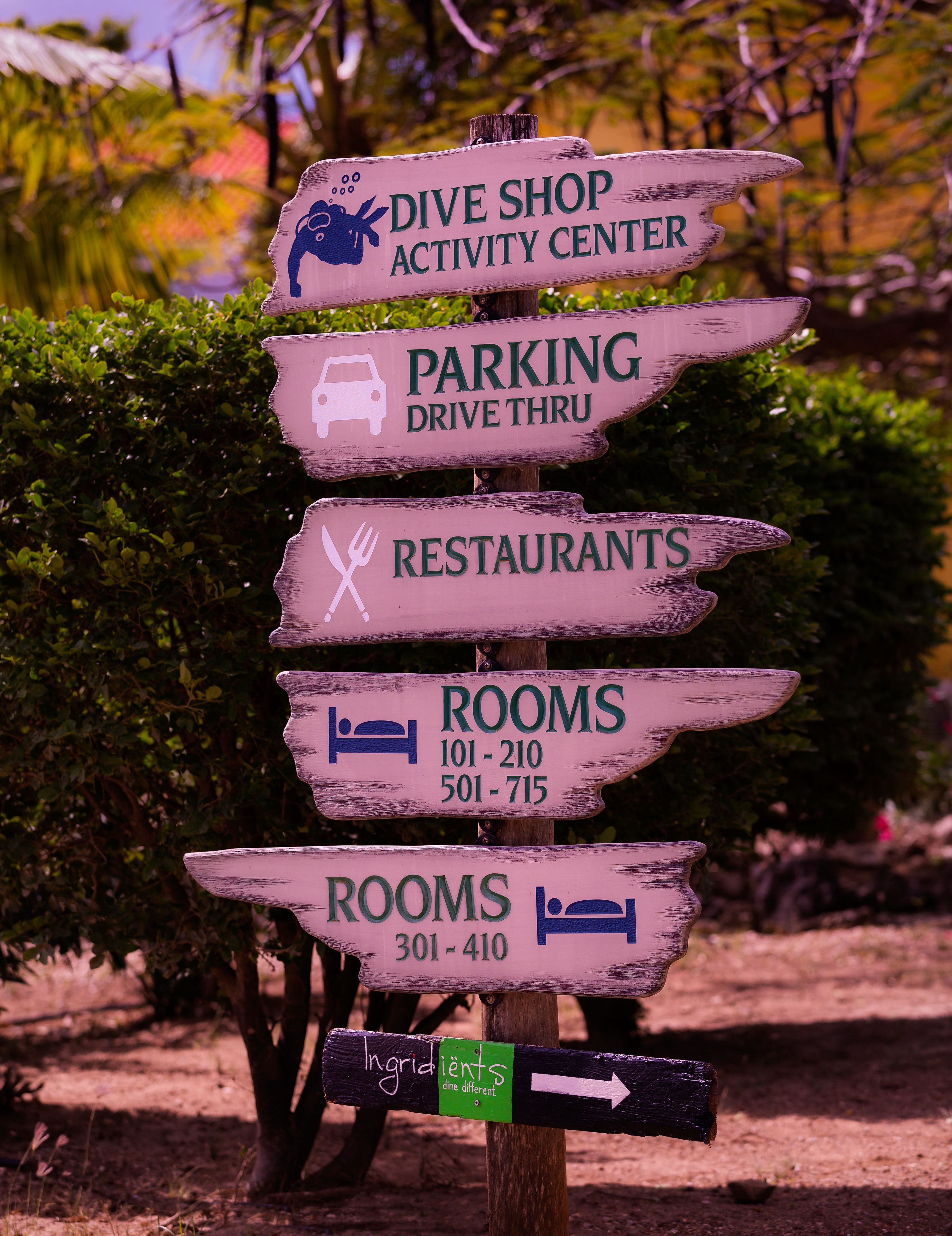 Ein Hinweisschild mit mehreren Pfeilen, die zu Einrichtungen wie einem Tauchgeschäft, Parkplätzen, Restaurants und Zimmern mit Kontaktdaten weisen.