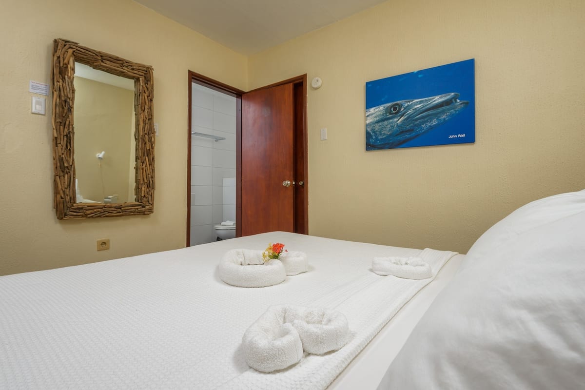A hotel room with a bed, a wooden-framed mirror, a painting of a whale, and a bathroom visible through an open door.