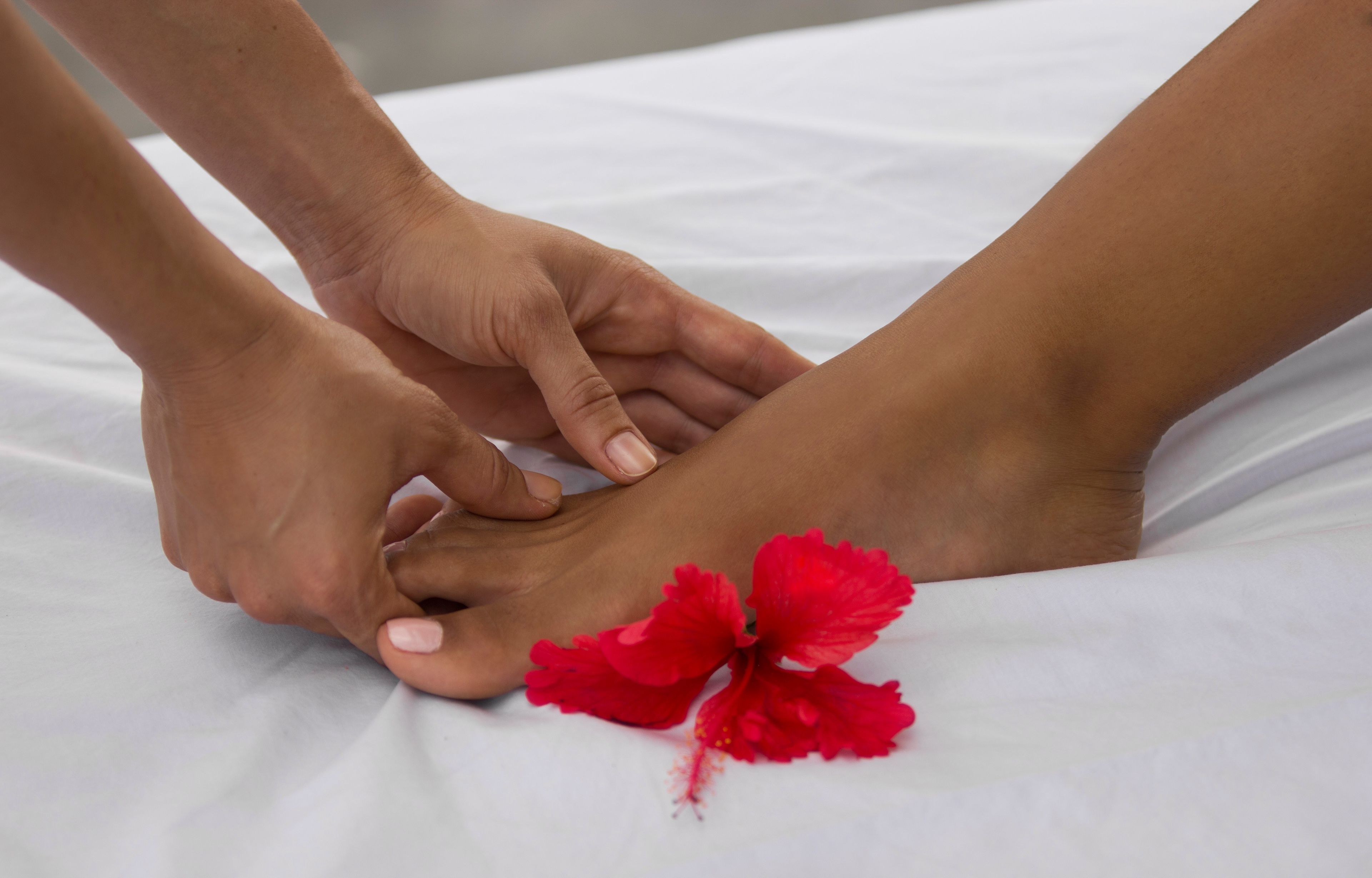 Primer plano de manos realizando un masaje de pies sobre una cama blanca con una flor de hibisco rojo.
