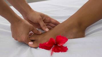 Primer plano de manos realizando un masaje de pies sobre una cama blanca con una flor de hibisco rojo.