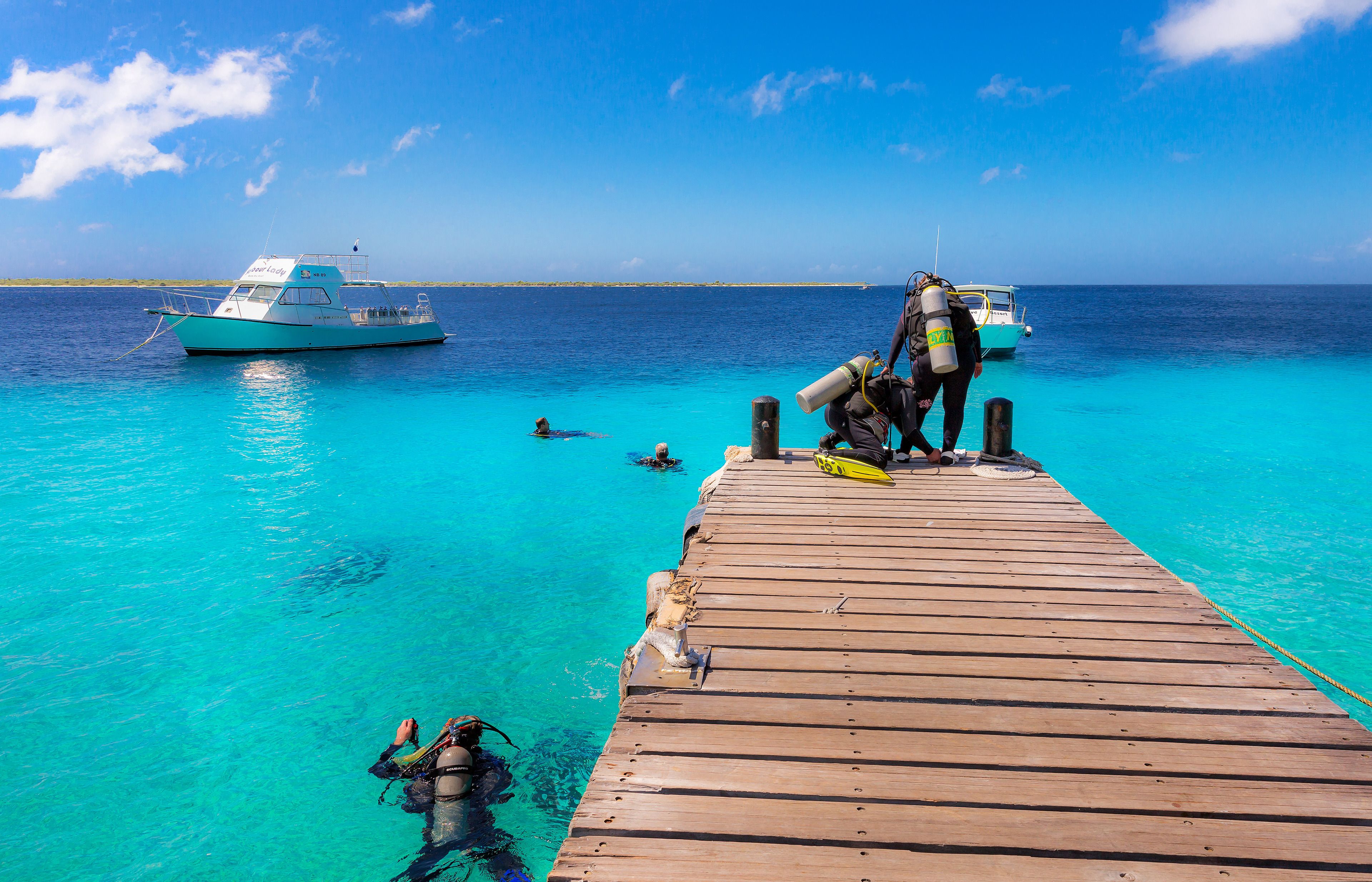 A wooden pier extending into clear turquoise water with divers and a boat in the background.