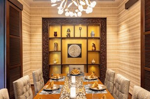 A dining area with a long table set for six, surrounded by upholstered chairs. The walls are adorned with patterned wallpaper and decorative shelves containing various items.