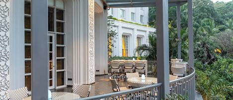 A covered outdoor seating area with wicker furniture and a glass ceiling, surrounded by lush greenery and a building in the background.