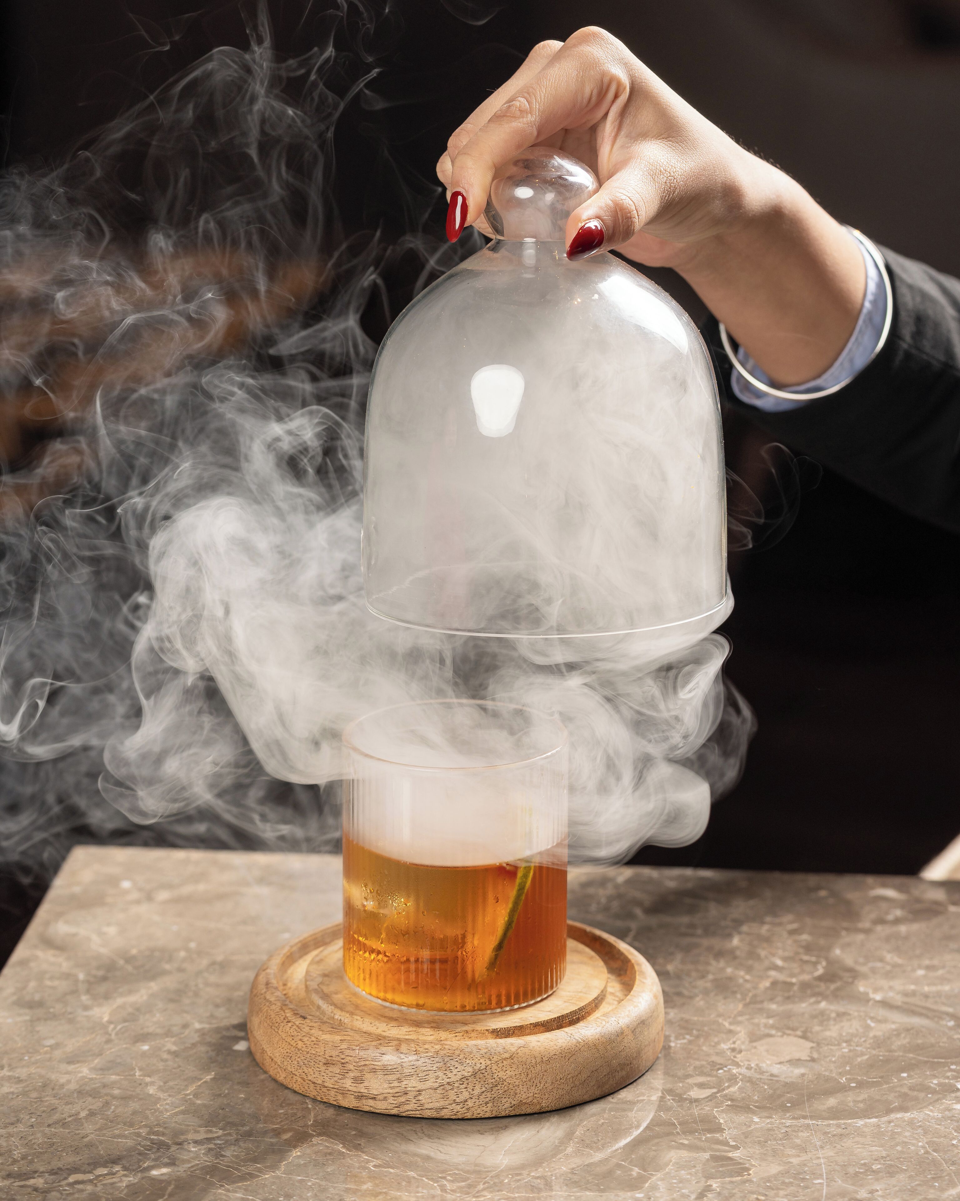 Una mano sostiene una cúpula de vidrio sobre un vaso con líquido ámbar, con vapor saliendo del vaso.