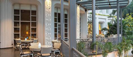 A covered outdoor seating area with tables and chairs, large windows, and a view of the surrounding greenery.