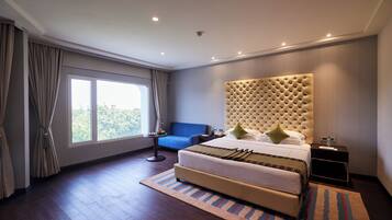 Un hotel moderno con una cama grande, un sillón azul, una mesita con una lámpara y vistas al exterior a través de un ventanal.