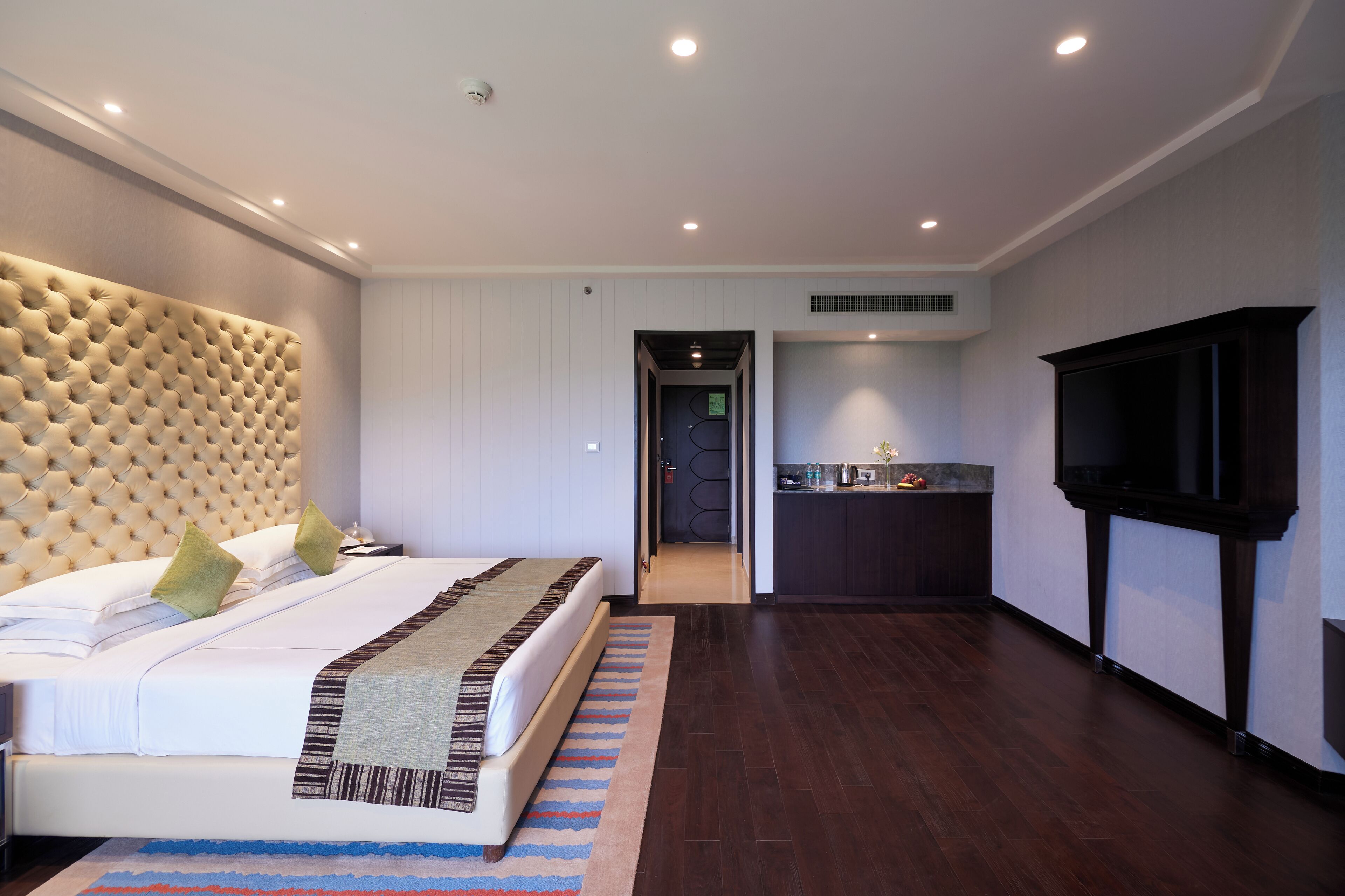 A modern hotel room with a large bed, a flat-screen TV, a small kitchenette, and a bathroom visible through an open door.