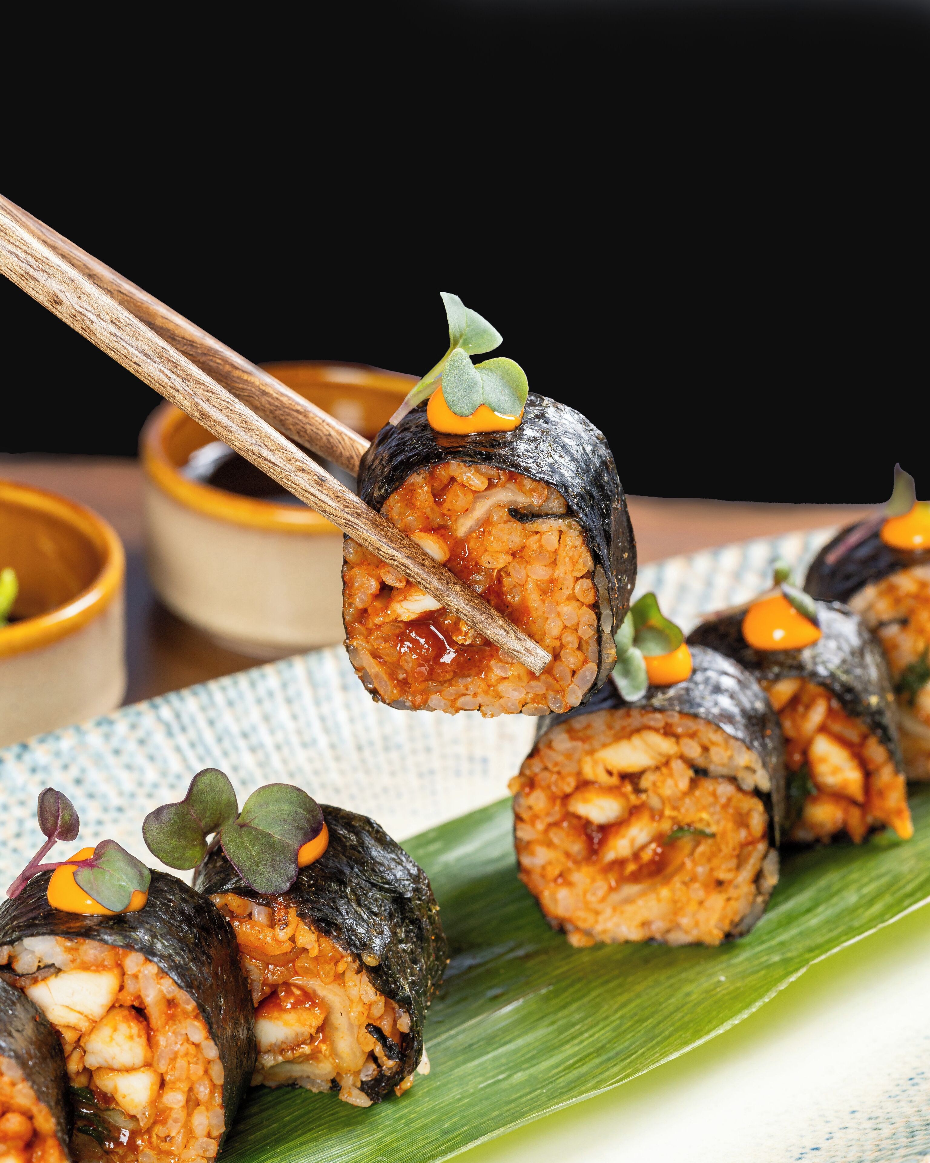 Des rouleaux de sushi servis avec des baguettes, décorés de feuilles et de tranches d’orange, présentés sur une assiette avec une feuille verte en dessous.
