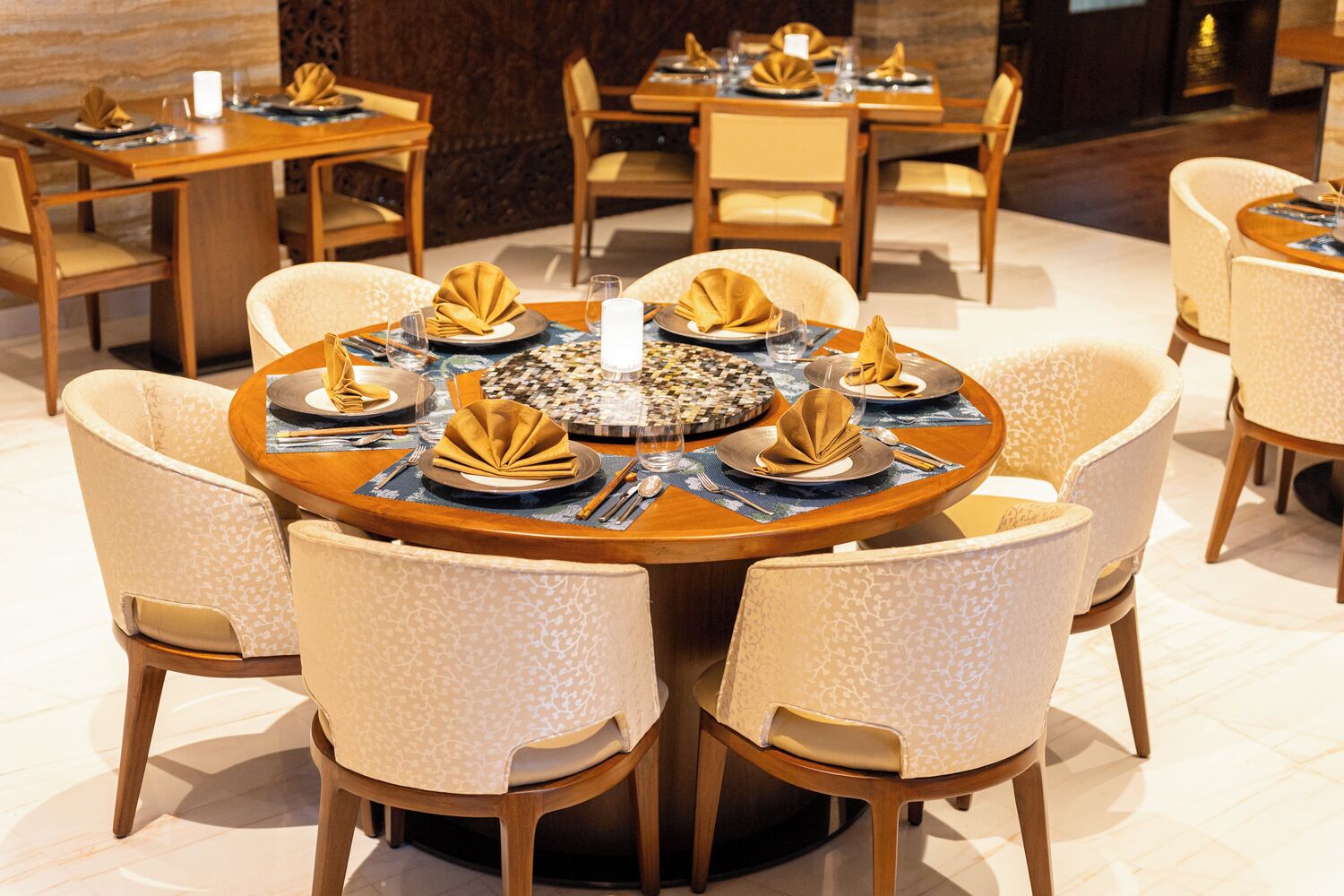 A dining area with round tables set for four, each with a white tablecloth, gold napkins, and a central candle.