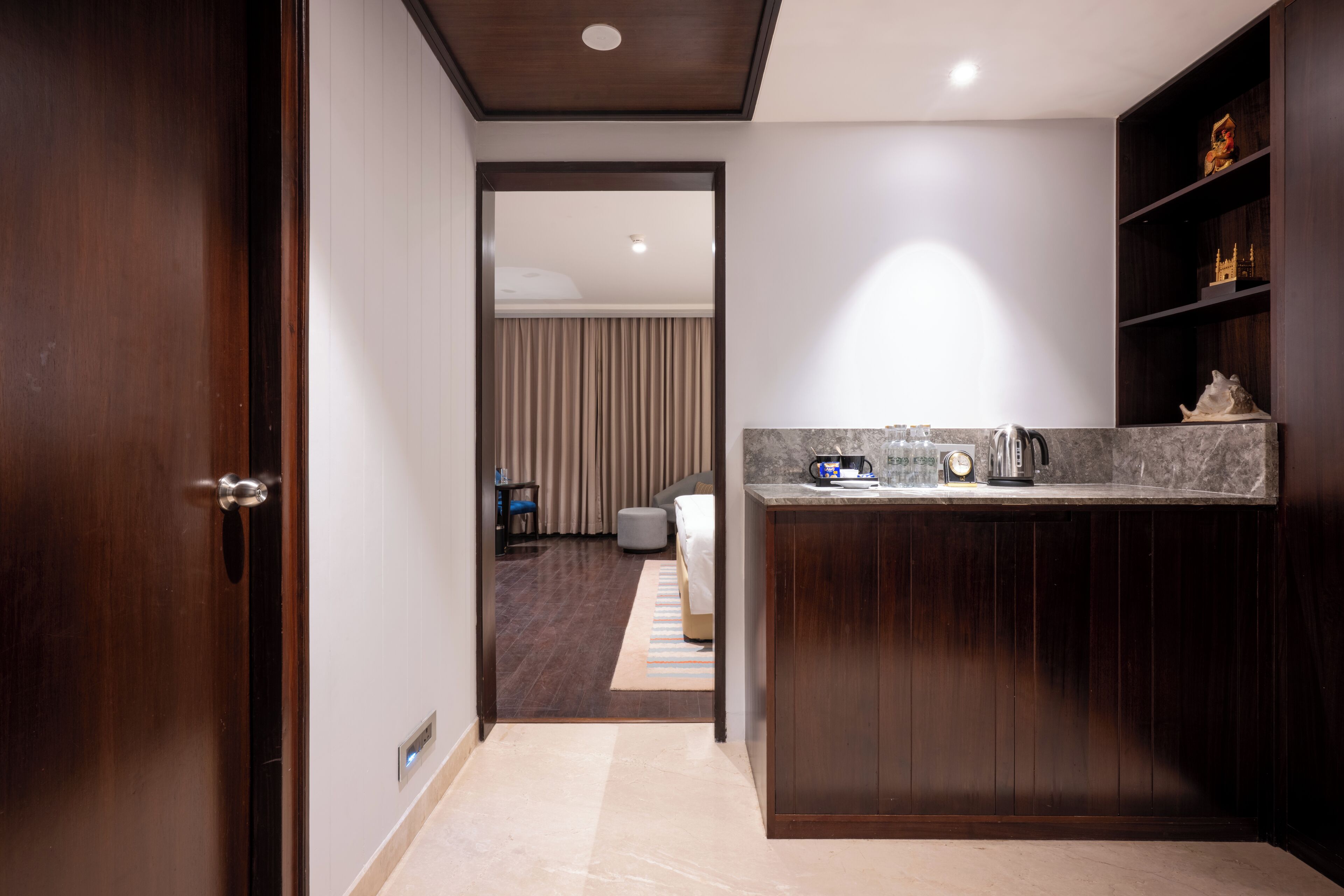 A modern hotel room with a wooden door, a kitchenette with a marble countertop, and a view into the adjacent bedroom.
