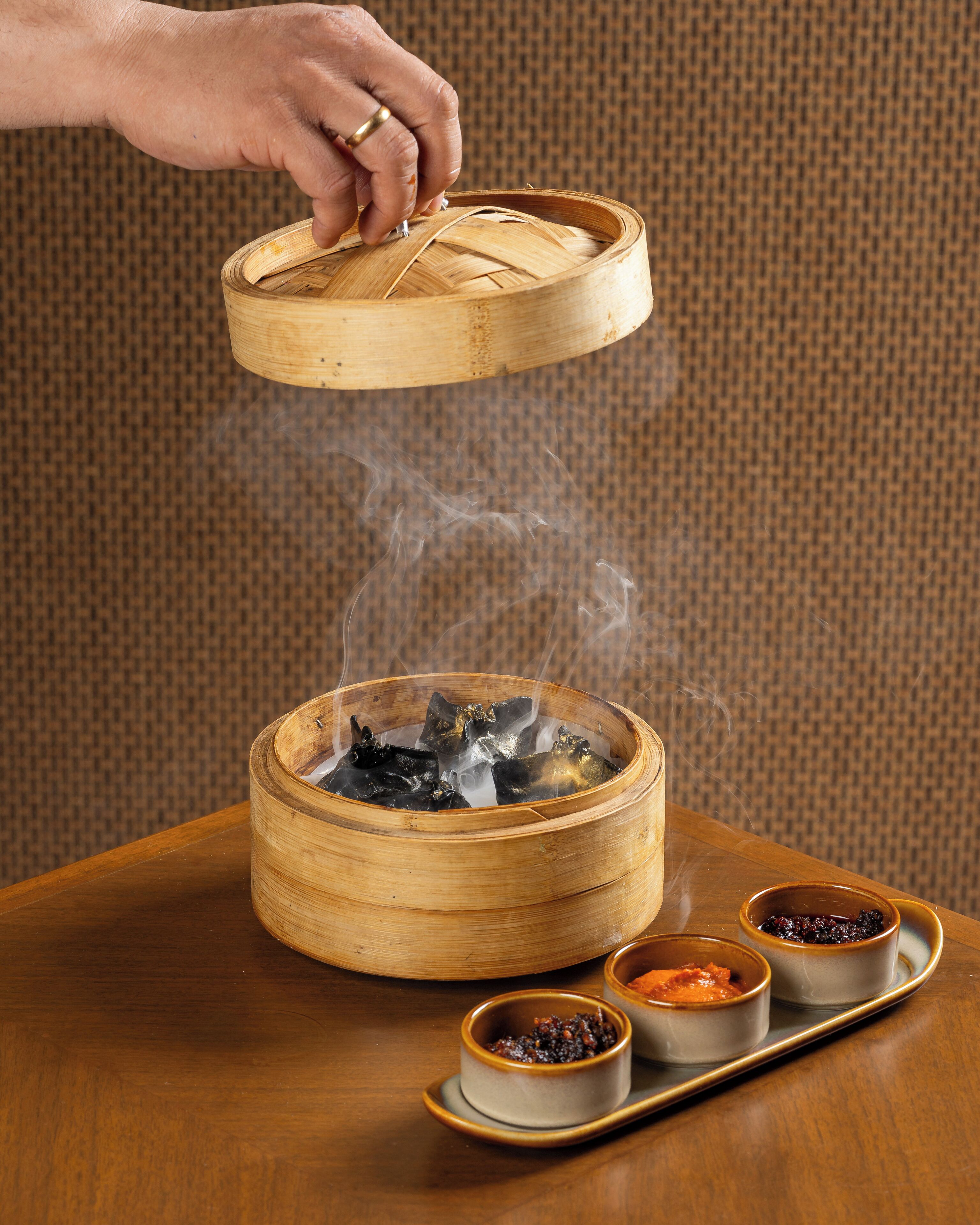 A hand lifting the lid of a bamboo steamer containing food, with steam rising. There are two small bowls with sauces on a tray beside the steamer.