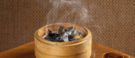 A hand lifting the lid of a bamboo steamer containing food, with steam rising. There are two small bowls with sauces on a tray beside the steamer.