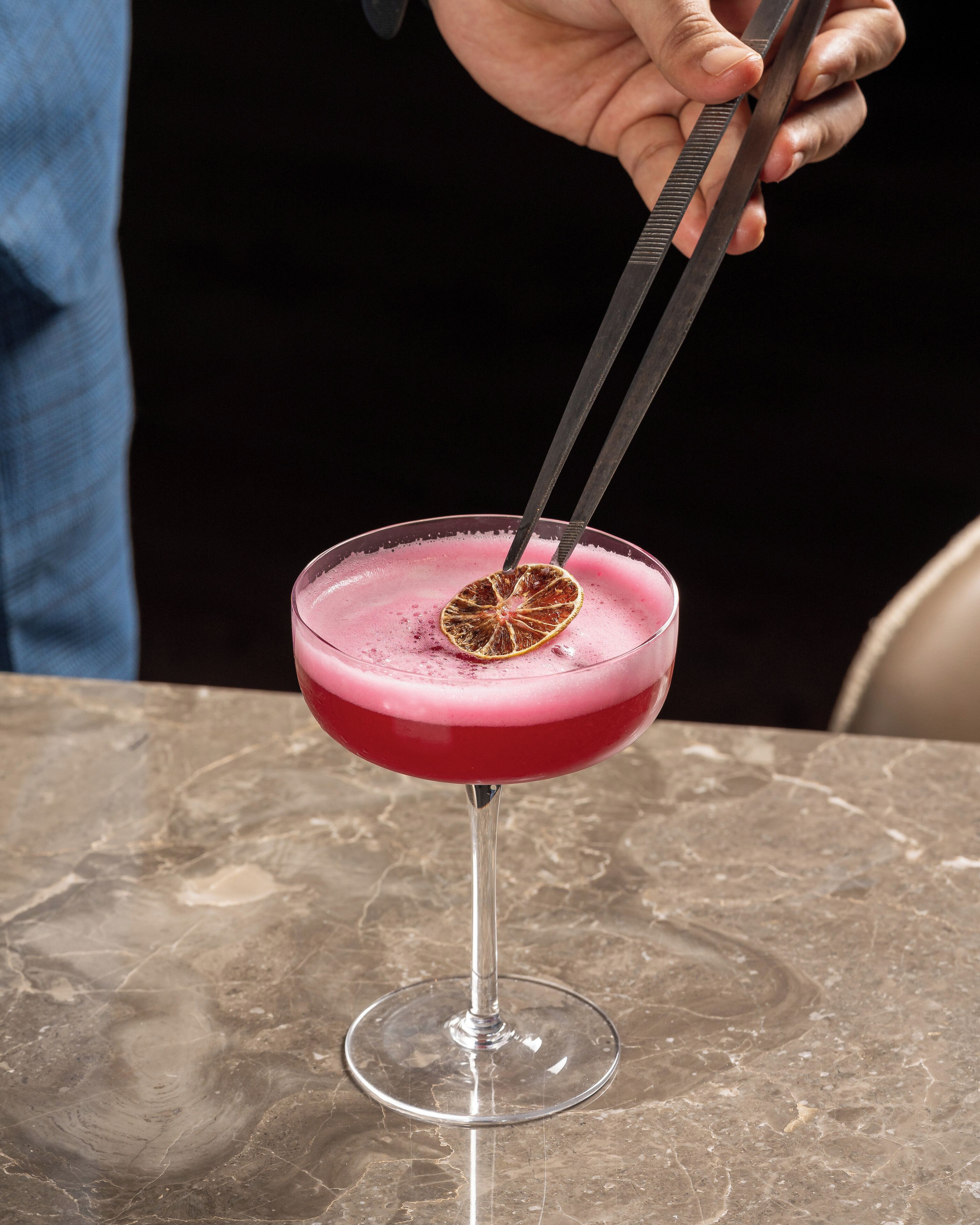 Un cocktail servi dans un verre, décoré d’une tranche d’orange séchée, que tient une personne à l’aide de pinces.