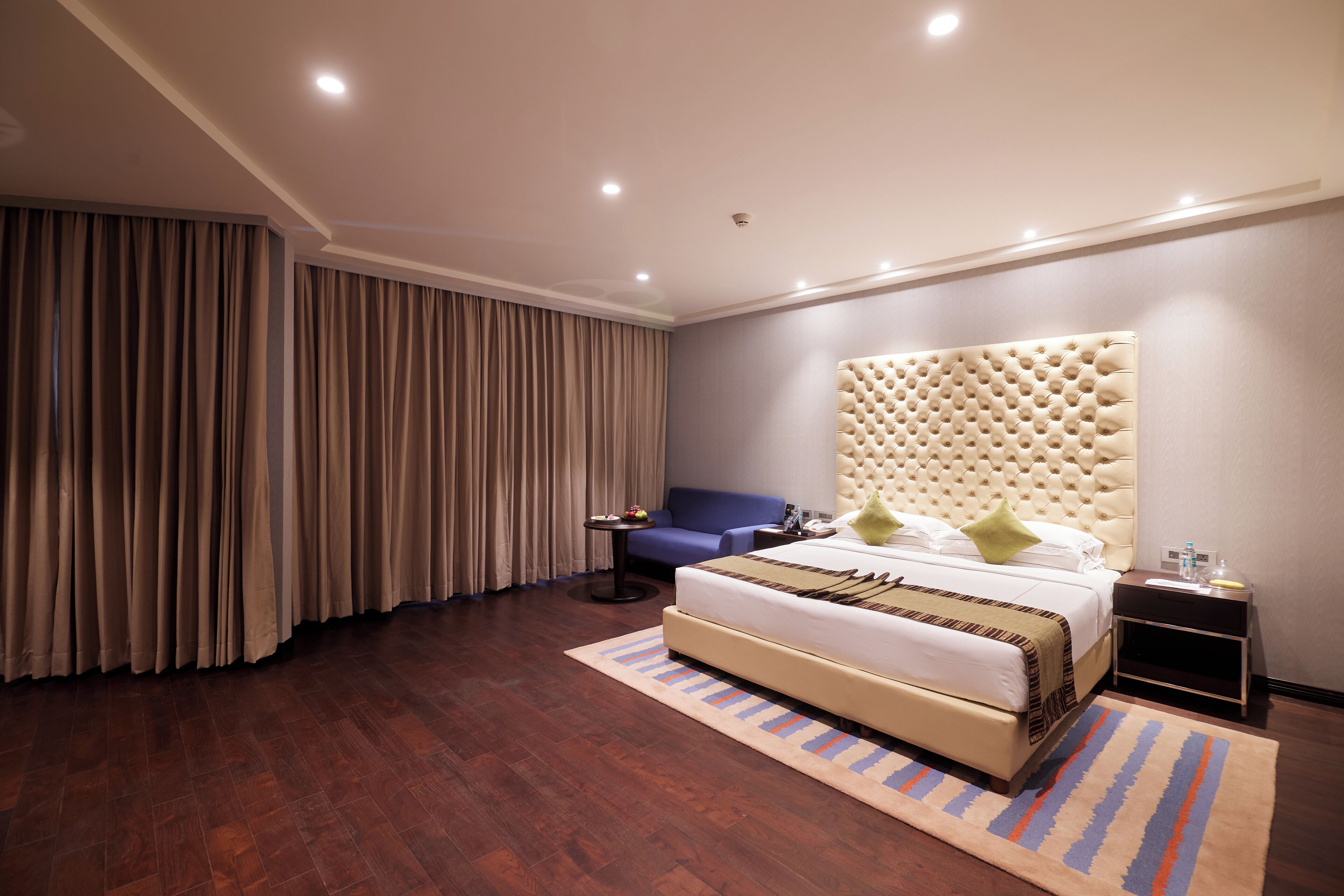 A modern hotel room with a large bed, a tufted headboard, a small side table, a blue armchair, and a large window with curtains.