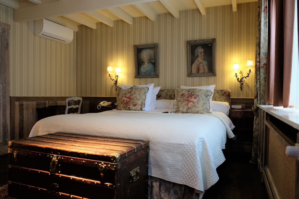 A hotel room with a large bed, floral-patterned pillows, two wall-mounted lamps, and a vintage suitcase.
