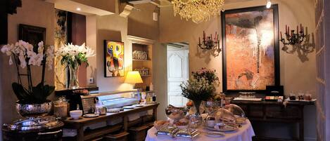 Un comedor con candelabro, mesa dispuesta con platos, vasos y flores. Hay un cuadro en la pared y una puerta que da a otra habitación.