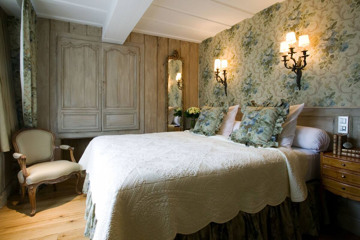 A bedroom with a large bed, a wooden wardrobe, a chair, and a nightstand with a lamp.